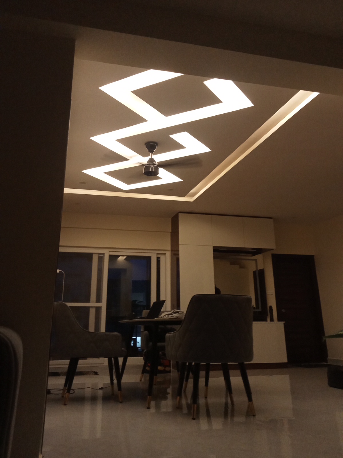 Dining area under a dramatic zigzag cove-lit false ceiling, with quilted grey dining chairs on brass-capped legs around a round table