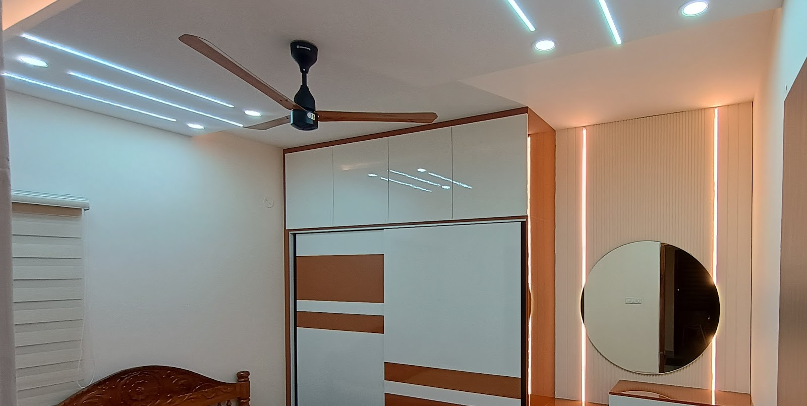 Bedroom corner with a white-and-tan sliding wardrobe under linear LED ceiling profiles, a carved wood bed edge in the foreground