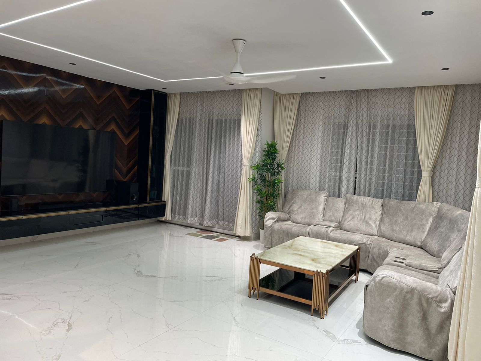 Living room with a chevron-pattern dark wood TV wall, a grey suede-style sectional sofa, and a marble-and-brass coffee table under a cove-lit ceiling.
