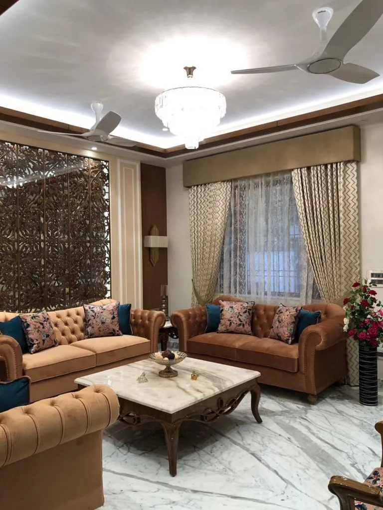 Living room with two tan velvet chesterfield sofas and floral-and-teal cushions around a carved-leg white marble coffee table