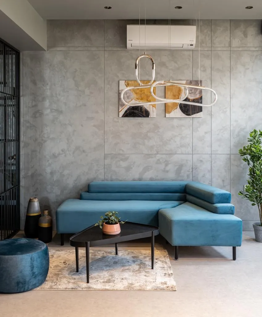 Teal velvet sectional set against a wall of grey concrete-finish slabs, with a looping brass-and-glass tube pendant hung across a two-panel ochre-and-black