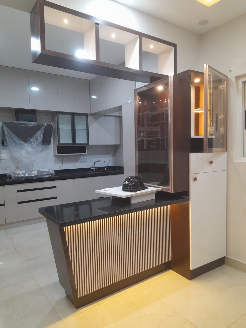 Kitchen counter with an adjoining glass-fronted pooja unit featuring a hanging brass bell and gold-lit interior.