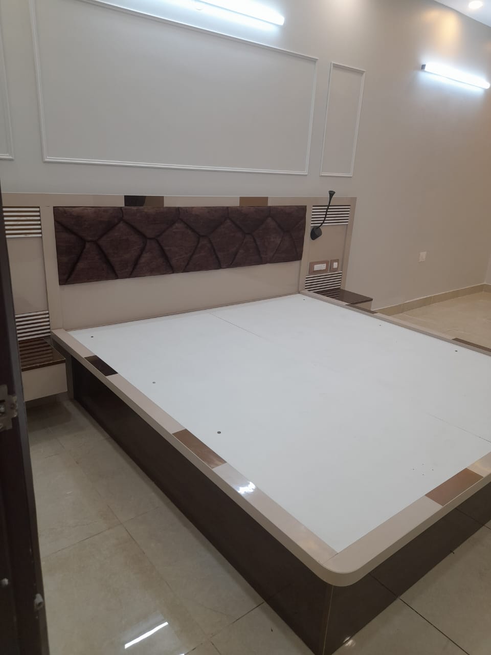 Real photo of a bedroom bed platform mid-handover with no mattress yet, a geometric tufted brown headboard, mirror-trimmed floating nightstands