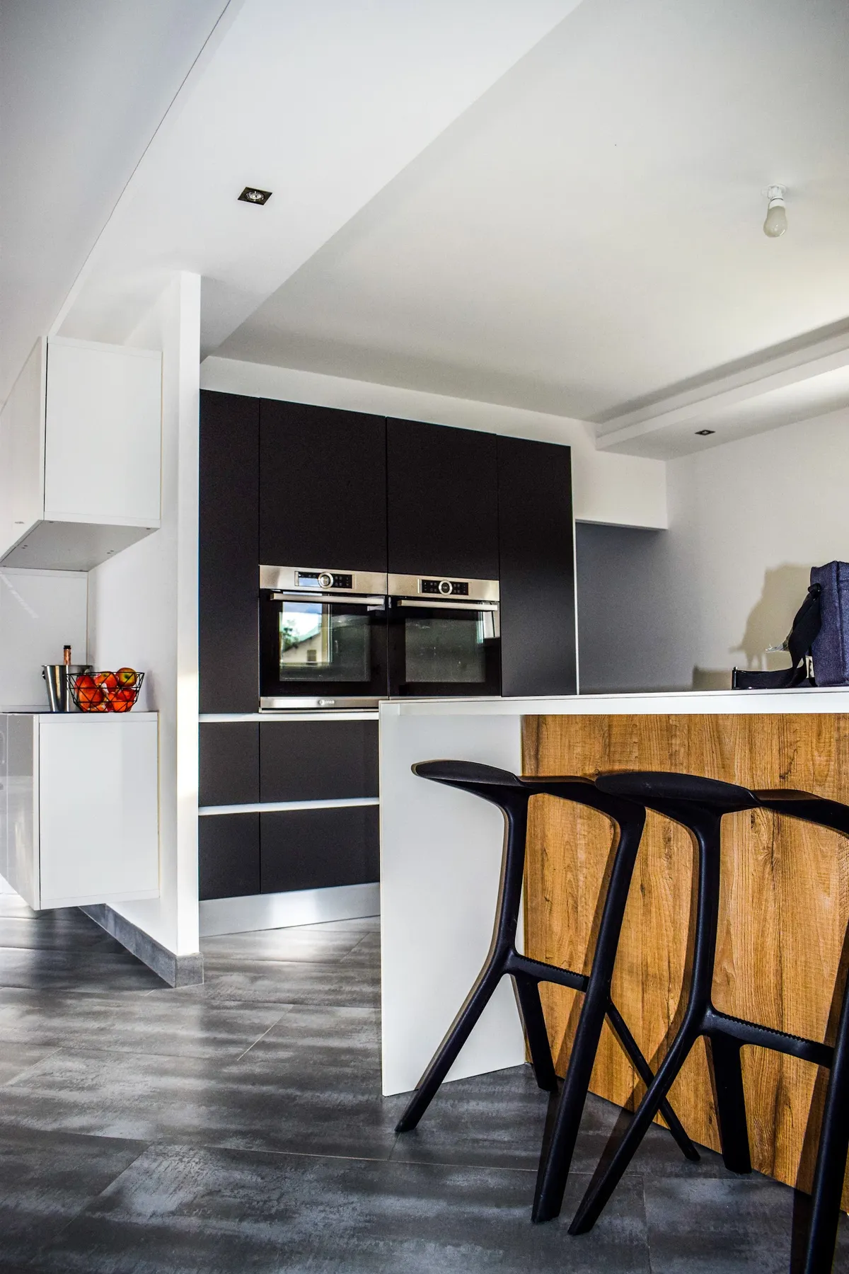 Black and white handleless kitchen tucked under a sloping stair soffit, with two stainless Bosch ovens stacked in the matte black tall bank and three black