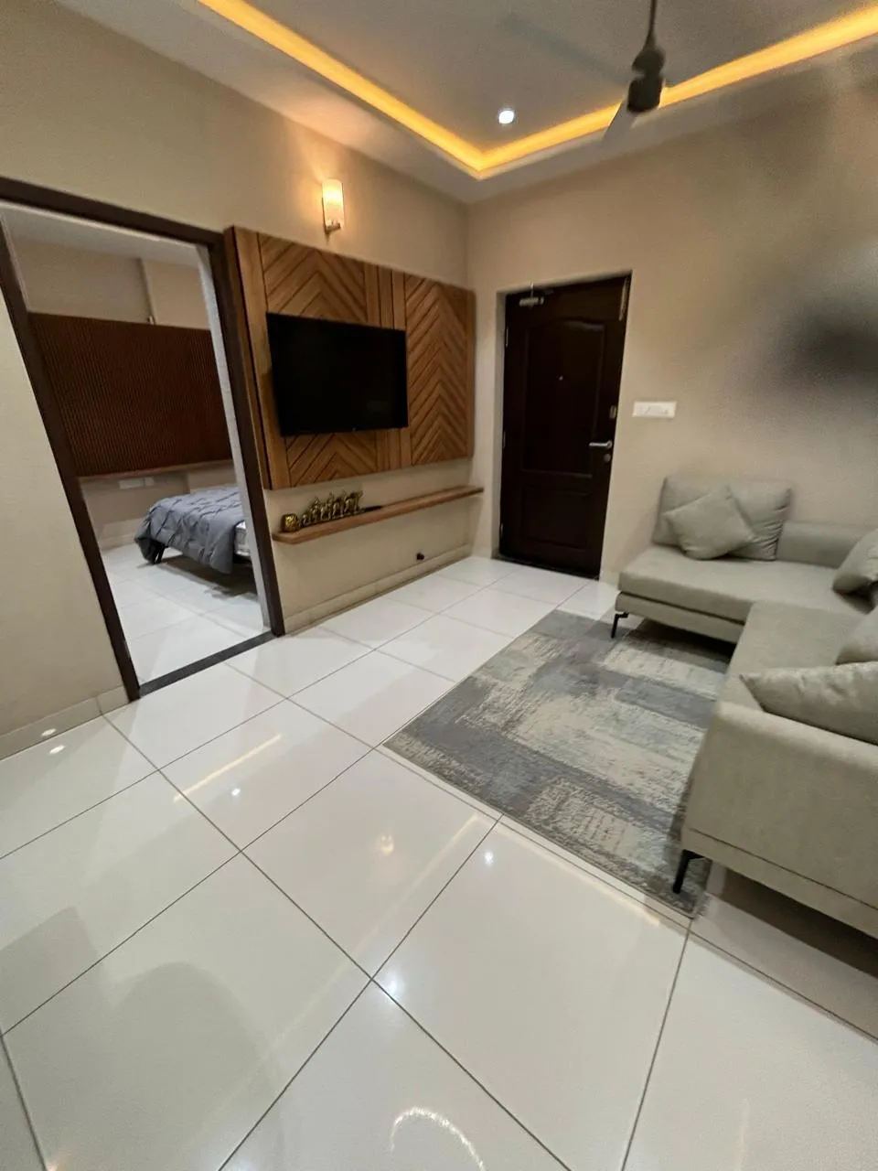 Compact flat living room where the TV sits on a chevron-laid teak panel with a slim brass-idol display ledge below, a grey L-sofa on a distressed grey rug