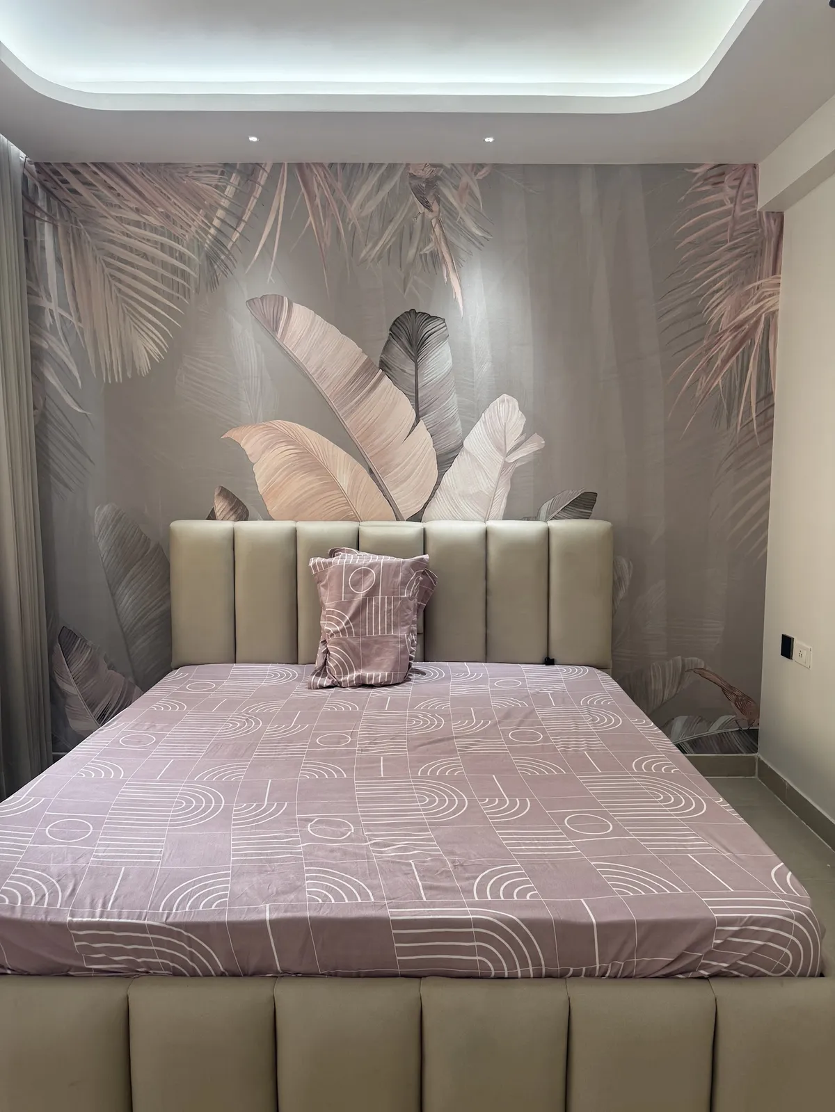 Bedroom with a blush tropical-leaf wallpaper mural filling the wall behind a wide cream channel-tufted headboard