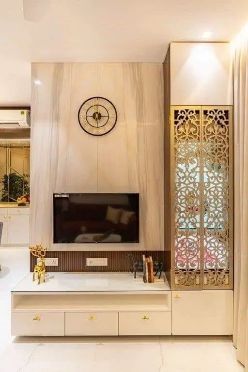 Living room TV wall in cream marble with a round wall clock, flanked by a tall cabinet with a gold jaali-pattern glass door, over a white floating TV console