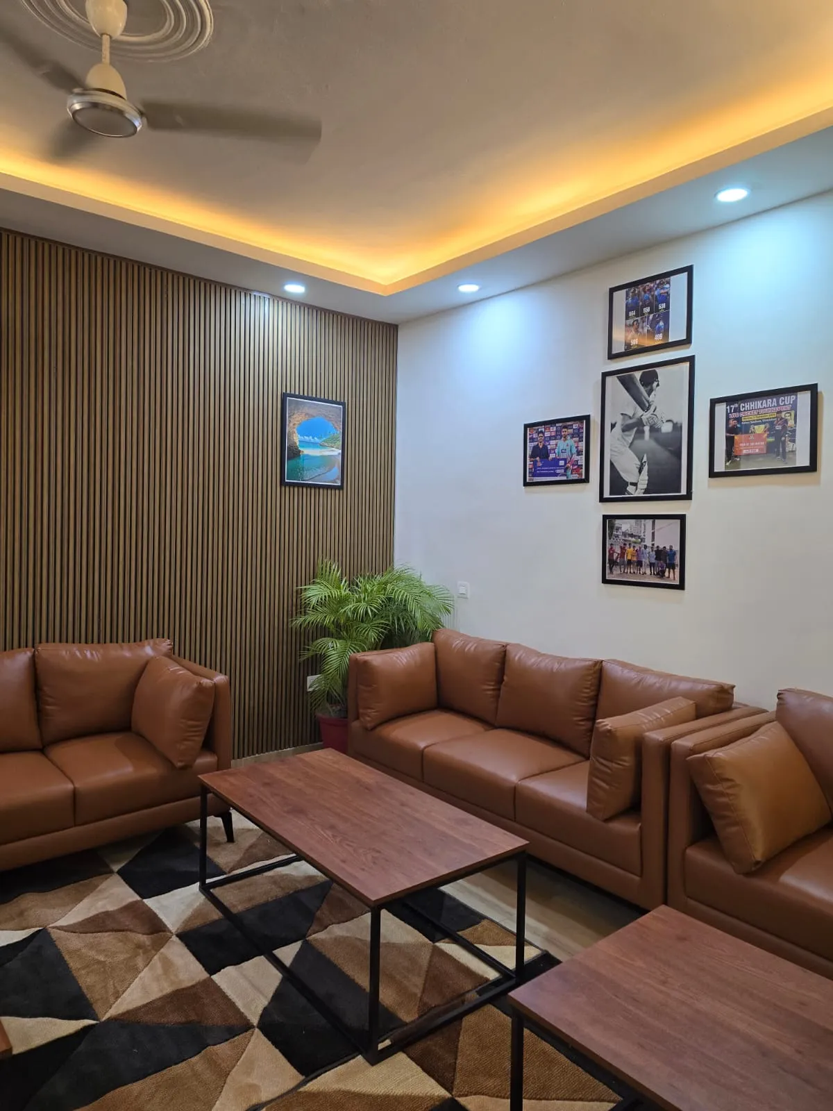 Tan leather 3+2 seater set against a full-height wooden slat wall, with a five-frame cricket photo gallery on the adjacent white wall and a black-and-tan