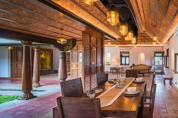 Traditional Kerala-style dining hall with a carved wood ceiling, amber glass pendant clusters, carved wood columns, and a long dark wood dining table.