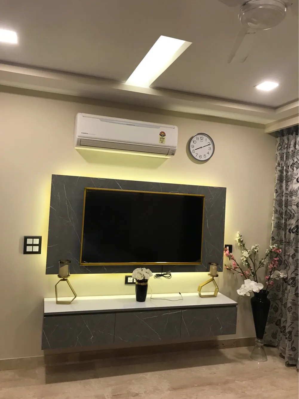 Living room TV unit with a backlit grey marble-effect panel in a gold frame, floating white console styled with gold candle stands and vases below