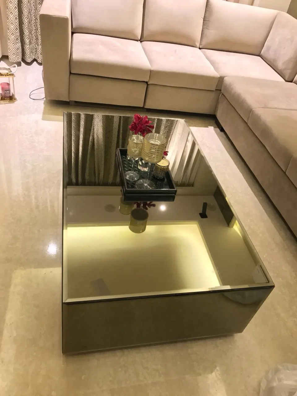 Mirror-clad rectangular coffee table with a backlit inner well and a black bar tray, shot from above against an L-shaped beige suede sectional on cream marble