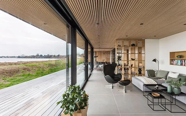 Long living room under a continuous oak-slat ceiling that runs out over the deck, with a black leather Egg chair and footstool facing a full-glass wall onto an