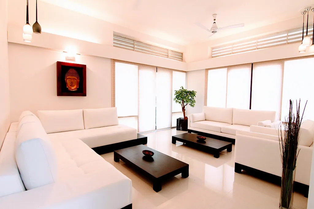All-white living room with two facing L-shaped sofas, dark wood coffee tables, a framed Buddha wall art and twin pendant lights over sheer-curtained windows