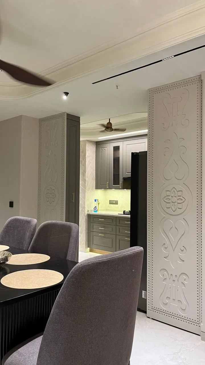 Dining area framed by tall carved relief-pattern wall panels, with a dark table and grey upholstered chairs opening into a grey shaker-style kitchen beyond