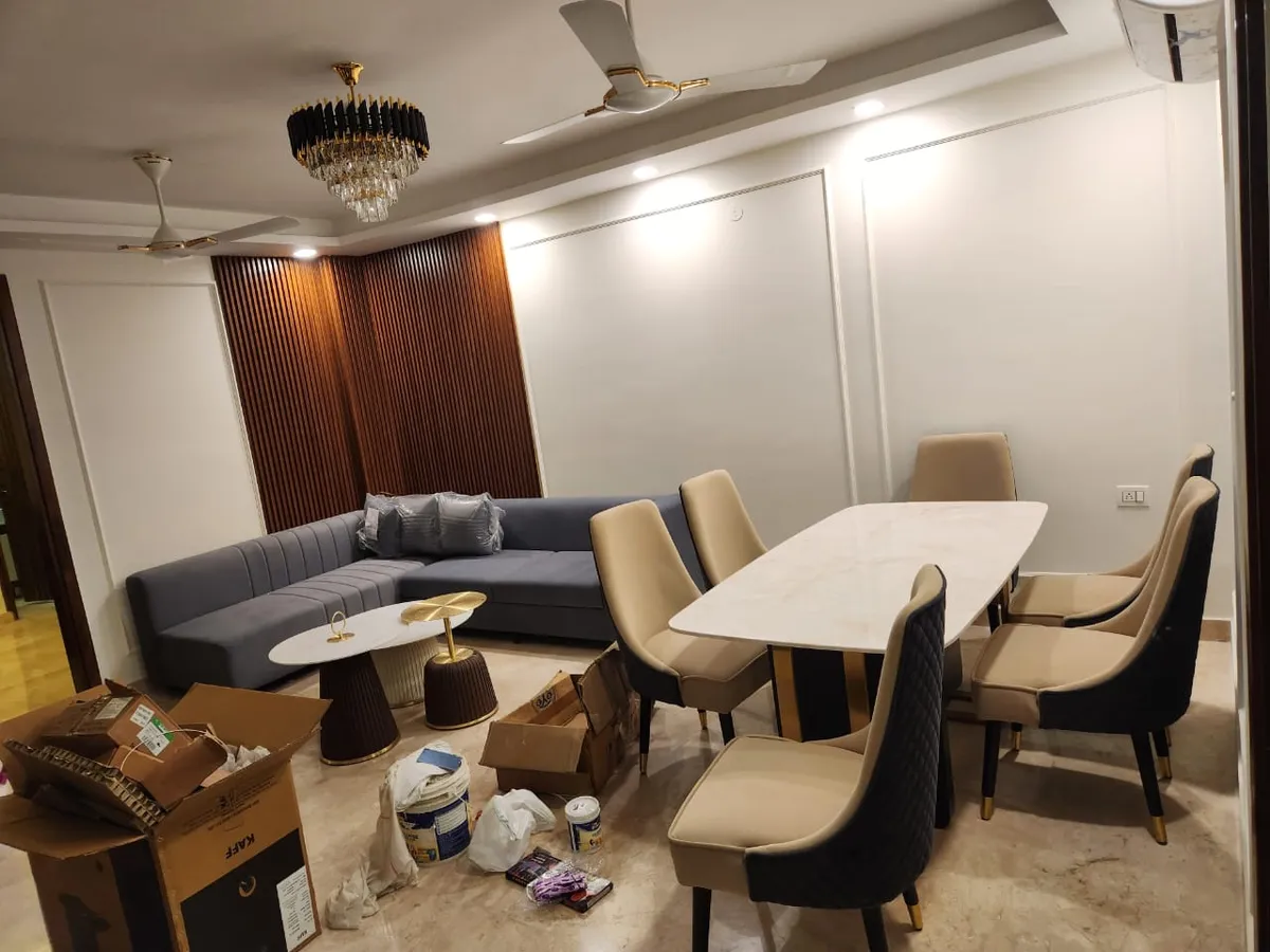 Moving-in day in an open living-dining: a grey channel-tufted L sofa still in its plastic wrap sits against a fluted teak panel wall under a black-and-crystal
