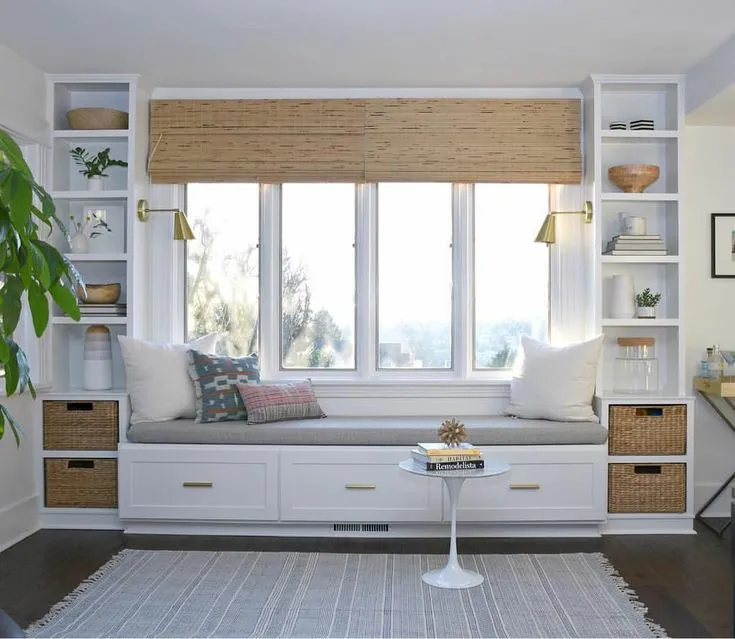 Built-in window seat nook with bamboo roman shades, brass reading sconces, flanking bookshelves with woven storage baskets and a round pedestal side table.