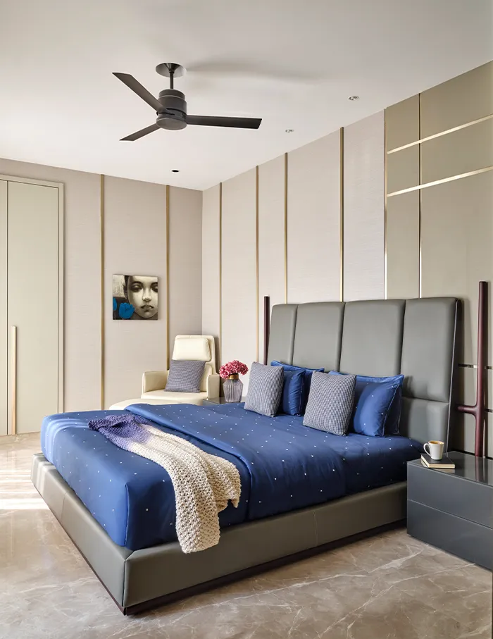 Master bedroom with a grey leather upholstered bed, navy dotted bedding, and a fluted wall panel with brass inlay strips behind a cream accent chair.