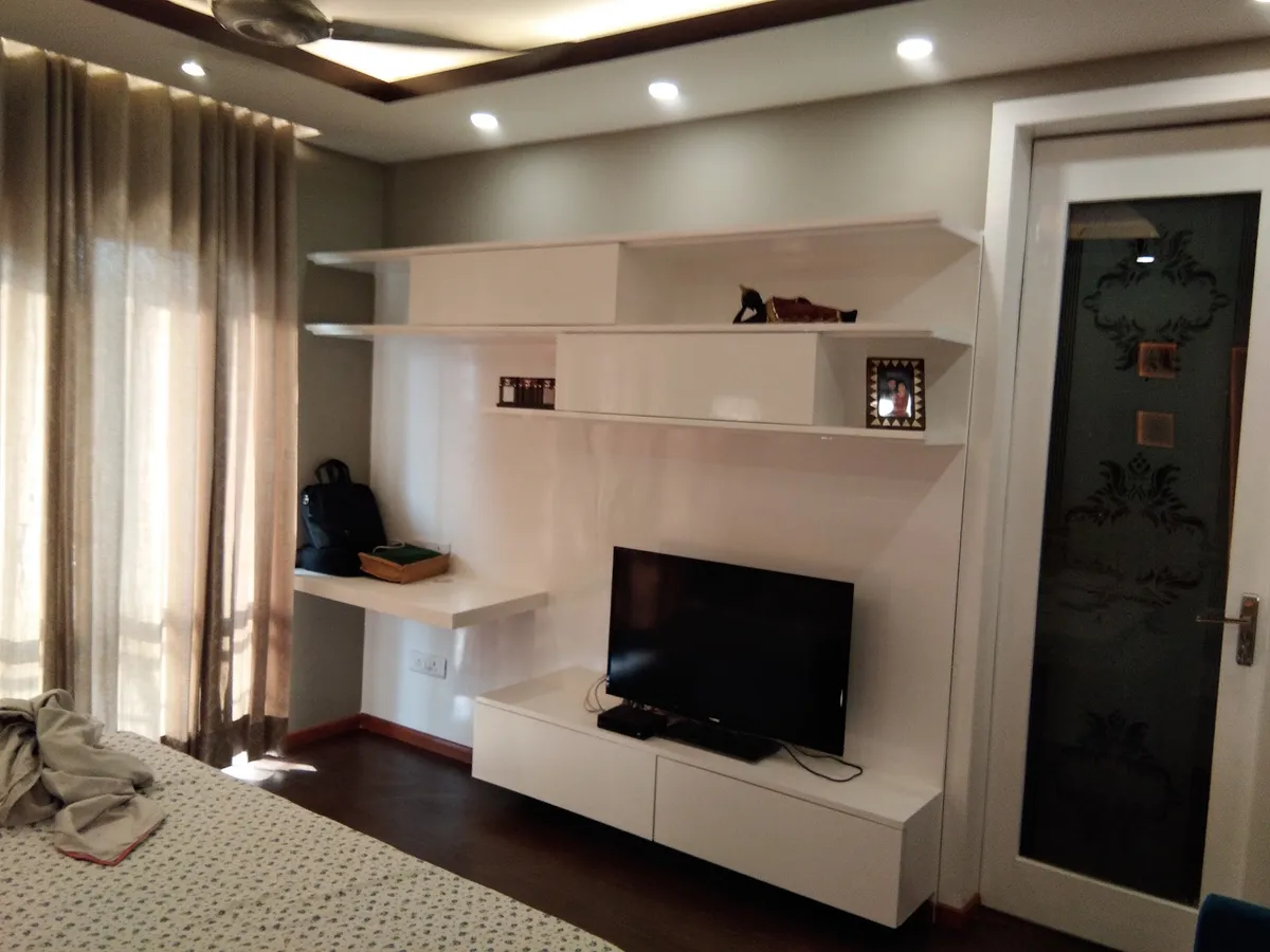 Bedroom TV wall in white lacquer - a wall-hung console with staggered open shelves and a small floating desk ledge to its left, beside an etched-glass door