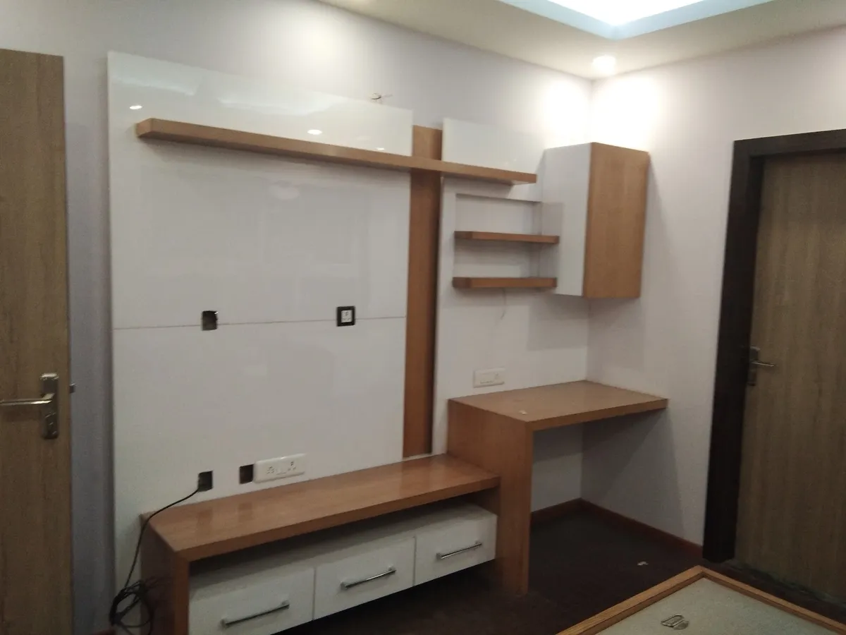 Bare study nook with a glossy white TV backdrop panel, wood floating shelves, and a built-in study desk beside a wall-mounted cabinet.