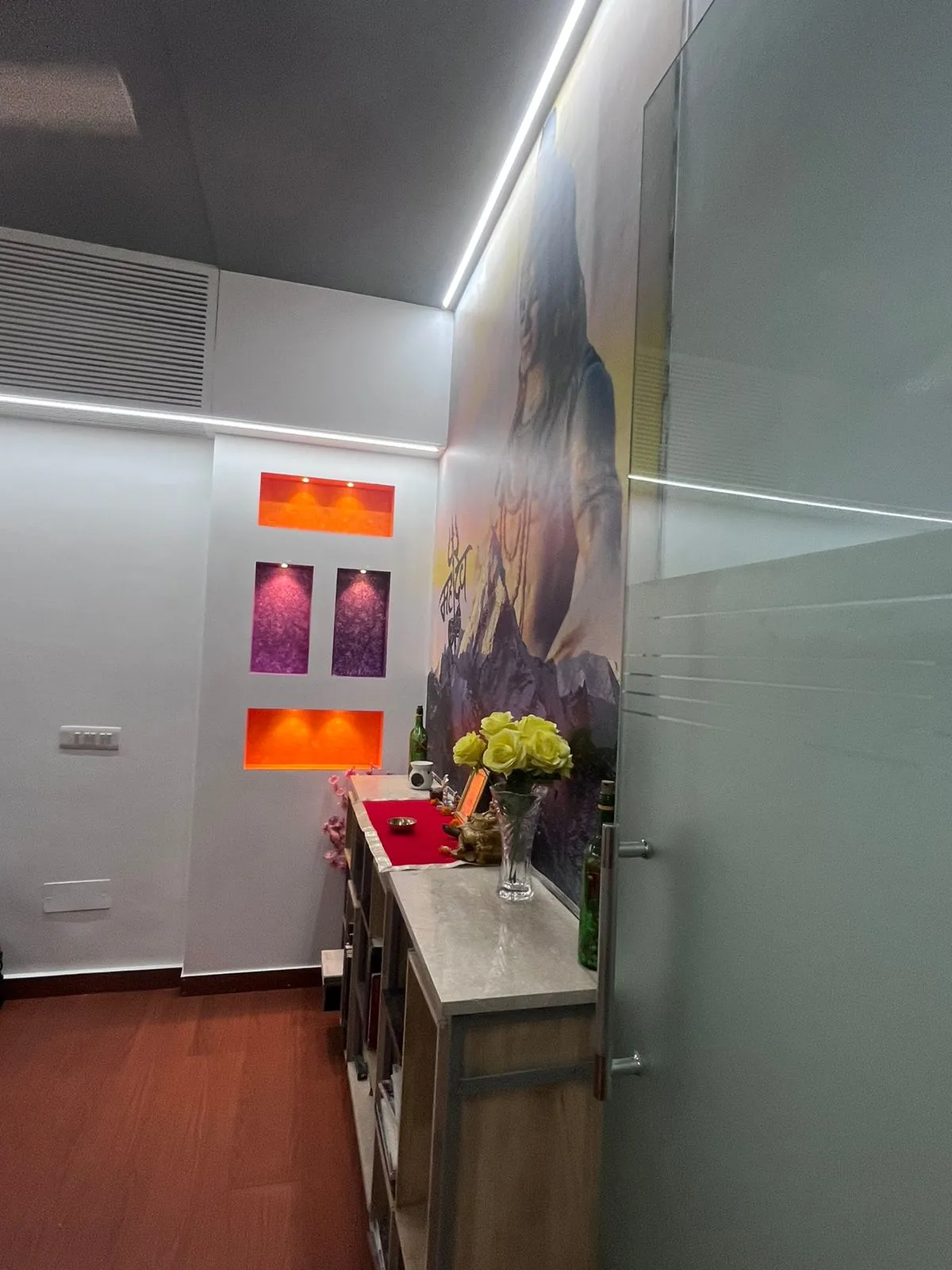 A backlit pooja corner in a hallway with a large printed mural of a deity, LED-lit orange and purple wall niches