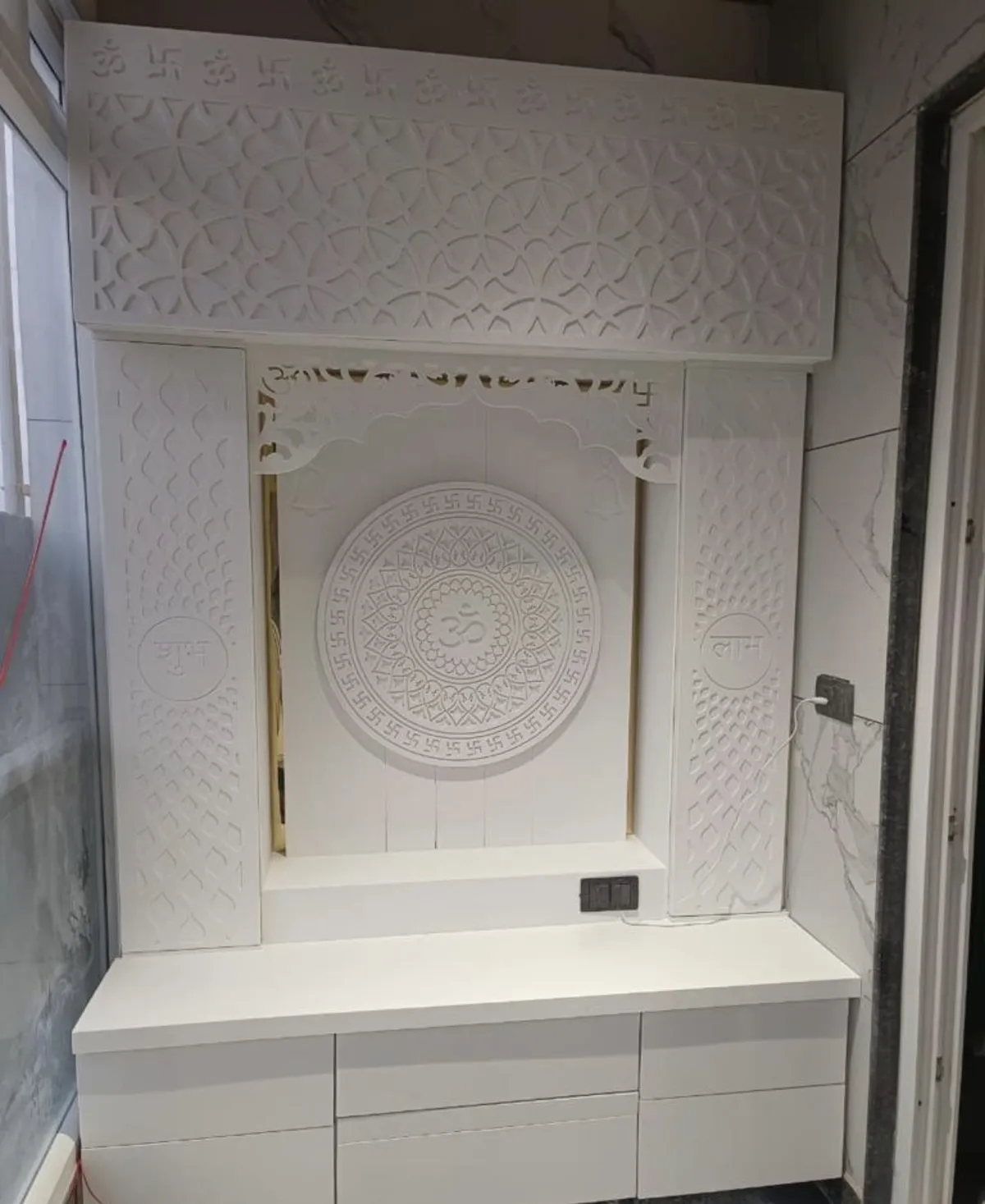 All-white CNC-carved mandir with an Om-and-swastik mandala back panel, Shubh and Labh carved pilasters, an Om-lettered jaali pelmet and a drawer bank below