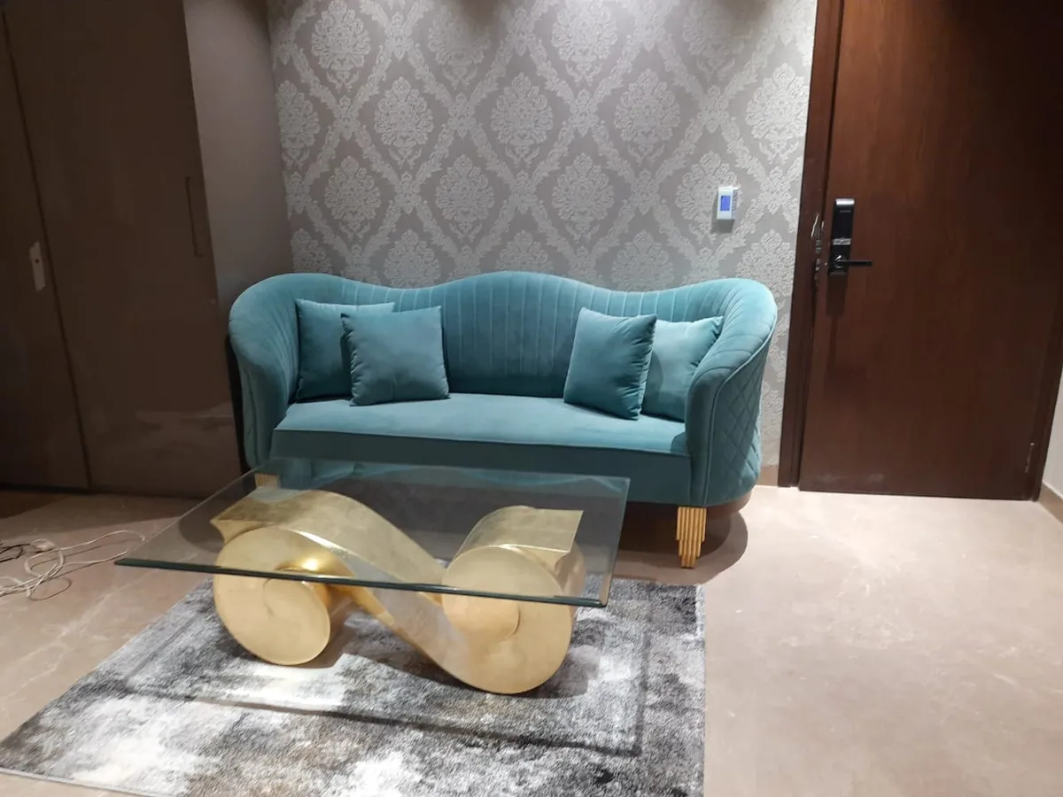 Seating nook with a teal channel-tufted velvet sofa against damask wallpaper, and a sculptural gold infinity-base glass coffee table on a grey rug.
