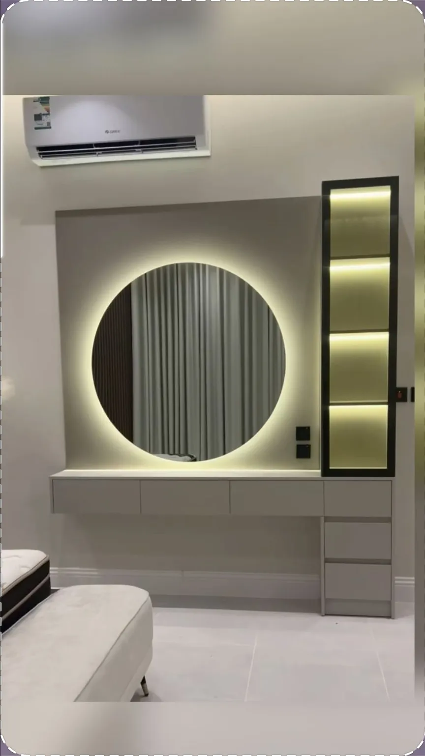Halo-lit round mirror over a wall-hung greige dressing counter with a drawer bank, next to a black-framed column of four warm-lit display niches