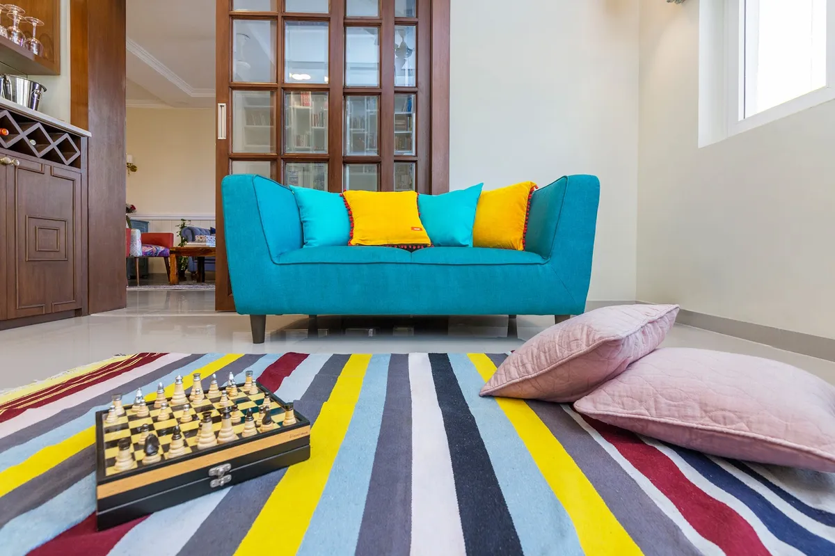 Living room with a teal sofa styled with yellow and turquoise cushions, a striped multicolor dhurrie rug with a chess-set vignette, pink floor cushions