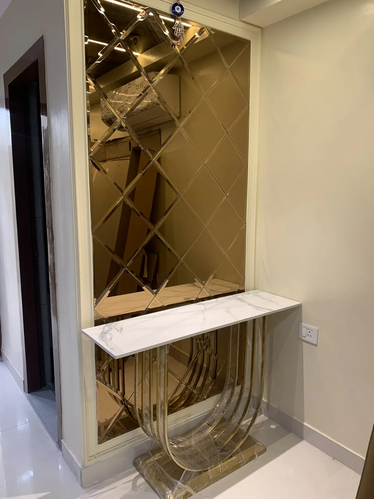 Entry console still in its shrink wrap — white marble top on a looping gold metal base against a full-height bronze-tinted bevelled diamond mirror panel