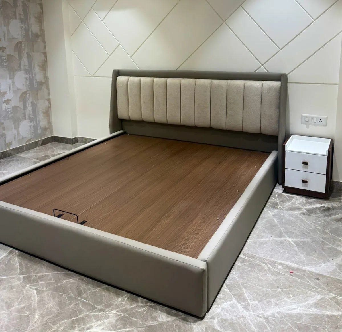 Bedroom under fit-out with a channel-tufted grey-and-beige storage bed against a herringbone wall panel, paired with a white two-drawer nightstand.