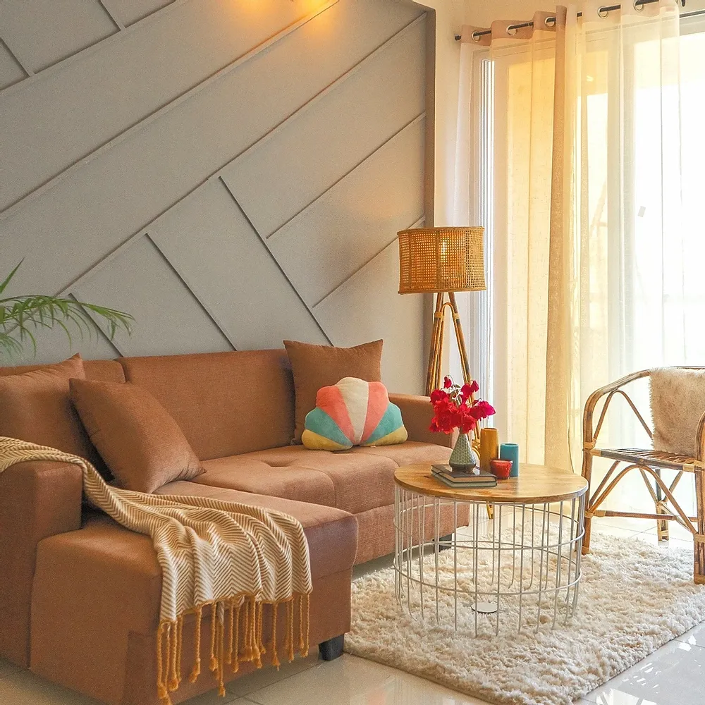Sunlit living room with a rust-brown L-sofa against a grey wall of diagonal moulded panels, a cane tripod floor lamp and rattan chair by sheer curtains
