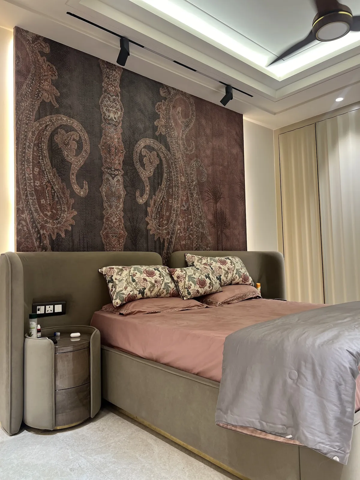 Master bedroom with a paisley wallpaper mural behind an upholstered wingback bed, dusty pink bedding and a matching round nightstand.
