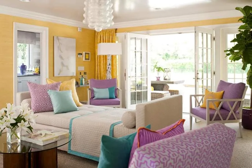 Sitting room with mustard yellow walls, a cushioned daybed in teal-trimmed white, purple armchairs, and French doors opening to a patio.