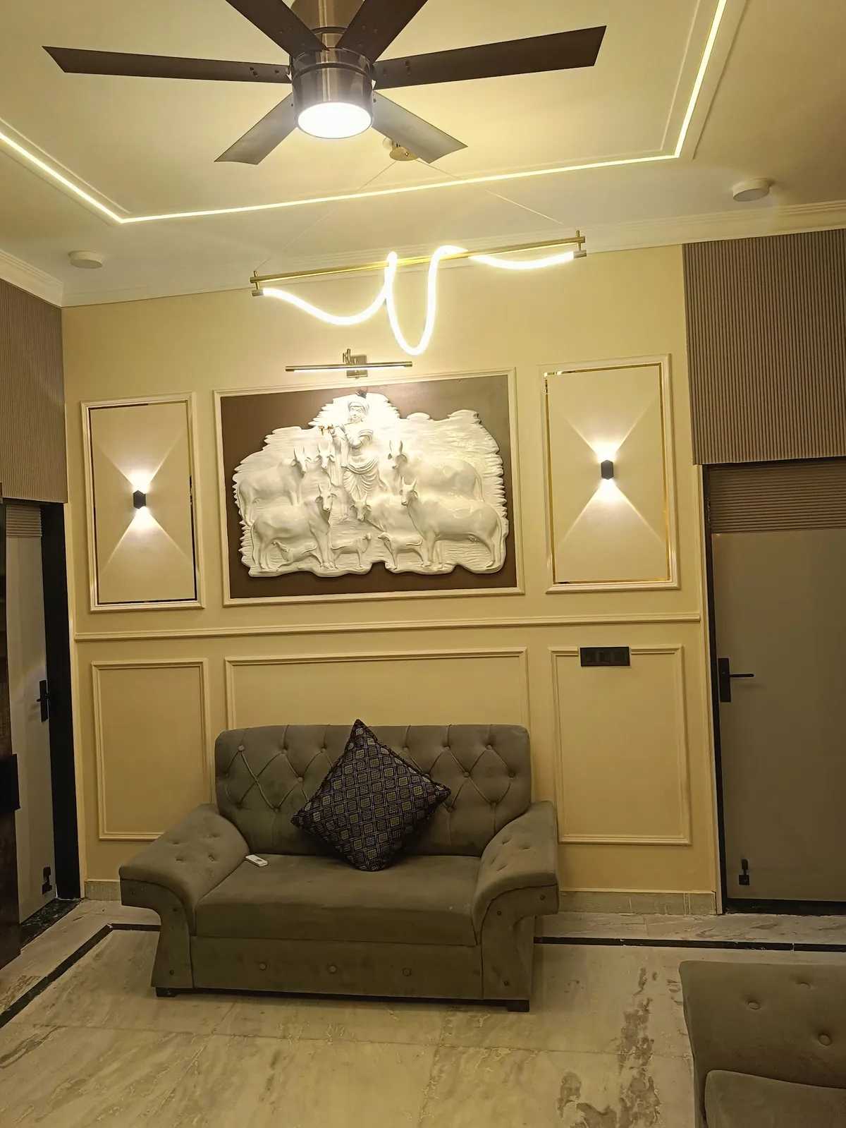 Living room with a carved relief sculpture of Krishna and cows set into a gold-trimmed moulded wall panel, flanked by wall sconces, above a tufted grey sofa.