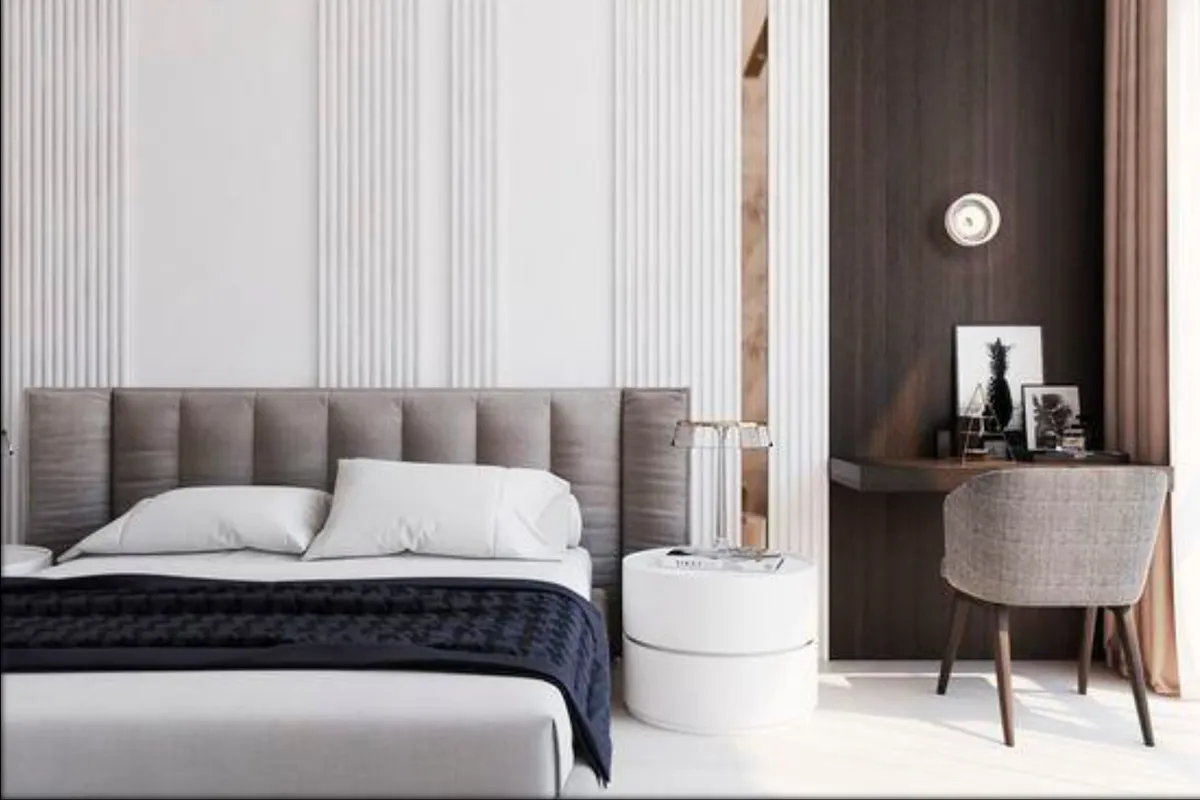 Render of a bedroom where a wide grey upholstered headboard runs across a white fluted wall panel, a round drum bedside table sits beside it