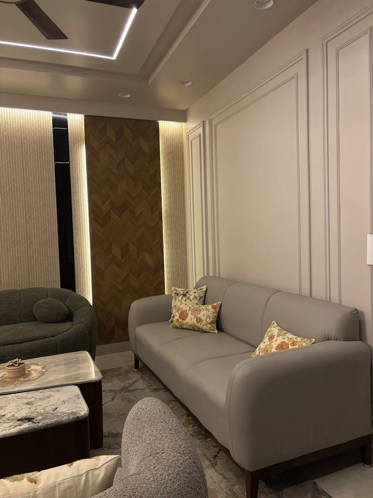 Living room with a grey sofa and green boucle armchair around nested marble coffee tables, a backlit chevron wood accent panel and moulded wall panelling.
