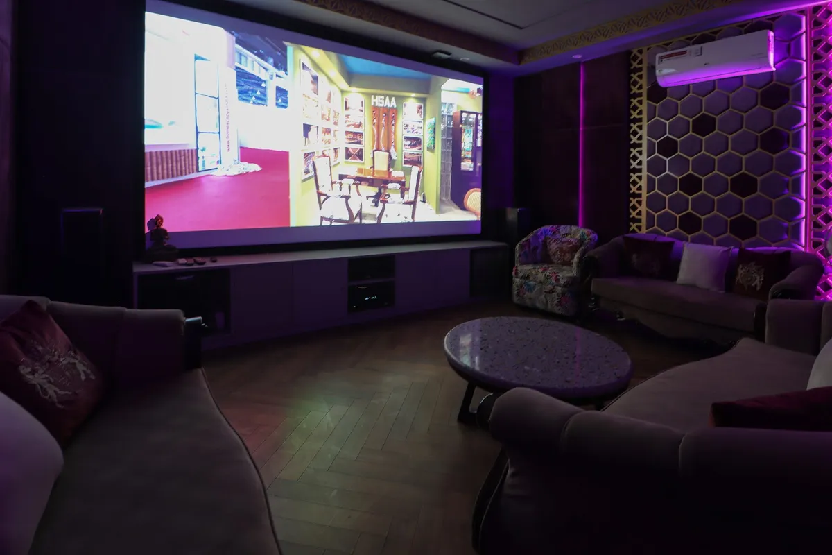 Darkened home theatre room with a wall-filling projection screen over a long low media console, plush taupe sofas and an oval stone coffee table