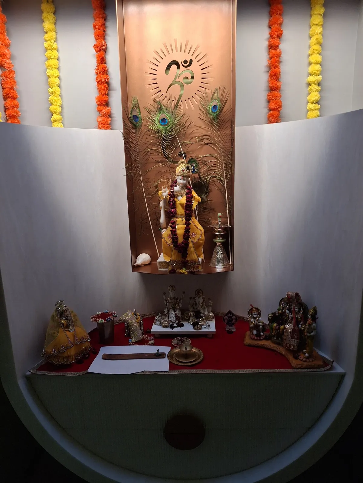 Built-in curved pooja niche with a backlit copper Om panel, peacock feathers and marigold garlands over a red-cloth altar shelf holding family deities.