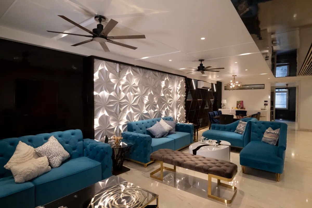 A living room with tufted teal velvet sofas and a gold-framed bench around a round marble coffee table, lit by a backlit 3D origami-pattern feature wall.