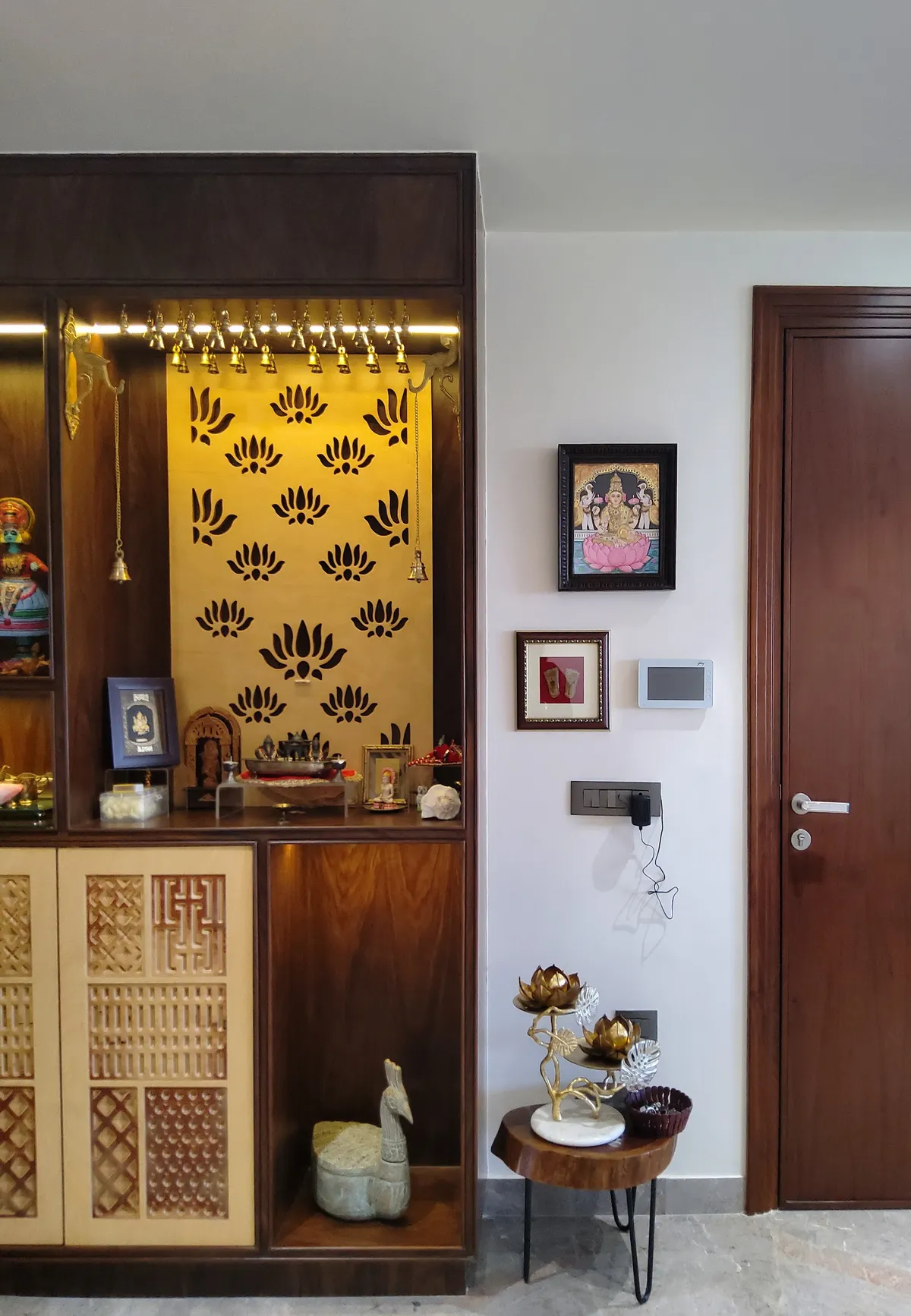 Teak-veneer mandir with a lotus-cut gold back panel and a rail of brass bells, next to a framed Tanjore Lakshmi and a live-edge side table holding brass lotus