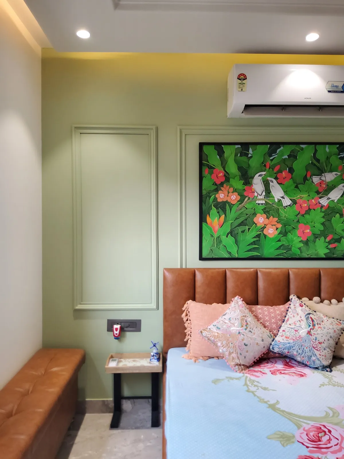 Sage-green bedroom wall picked out with slim applied mouldings, a Pichwai-style painting of mynahs among hibiscus hung over a tan channel-tufted leather
