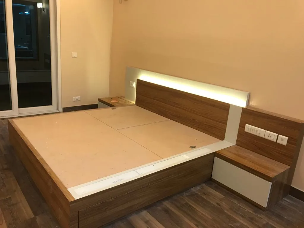 Bed under construction showing a wood-veneer platform frame with an integrated LED-backlit headboard and a floating attached nightstand.