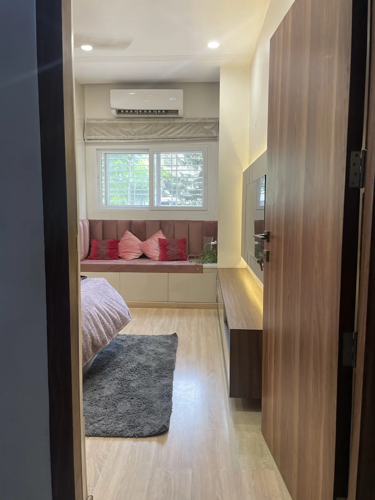 A bedroom viewed through the doorway, with a mauve channel-tufted window seat bench topped with pink and red cushions, a grey shag rug