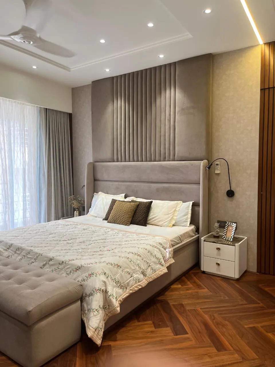Grey suede headboard wall carried floor-to-ceiling with a channel-tufted centre panel, teak herringbone floor