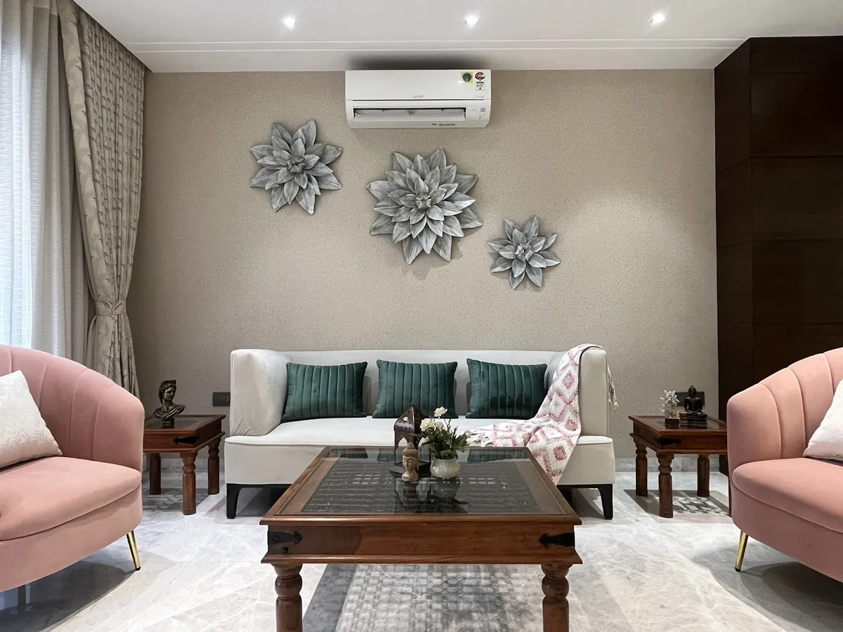 A living room with a cream sofa flanked by pink velvet armchairs, a carved wood coffee table, and a trio of oversized metal flower wall sculptures above.