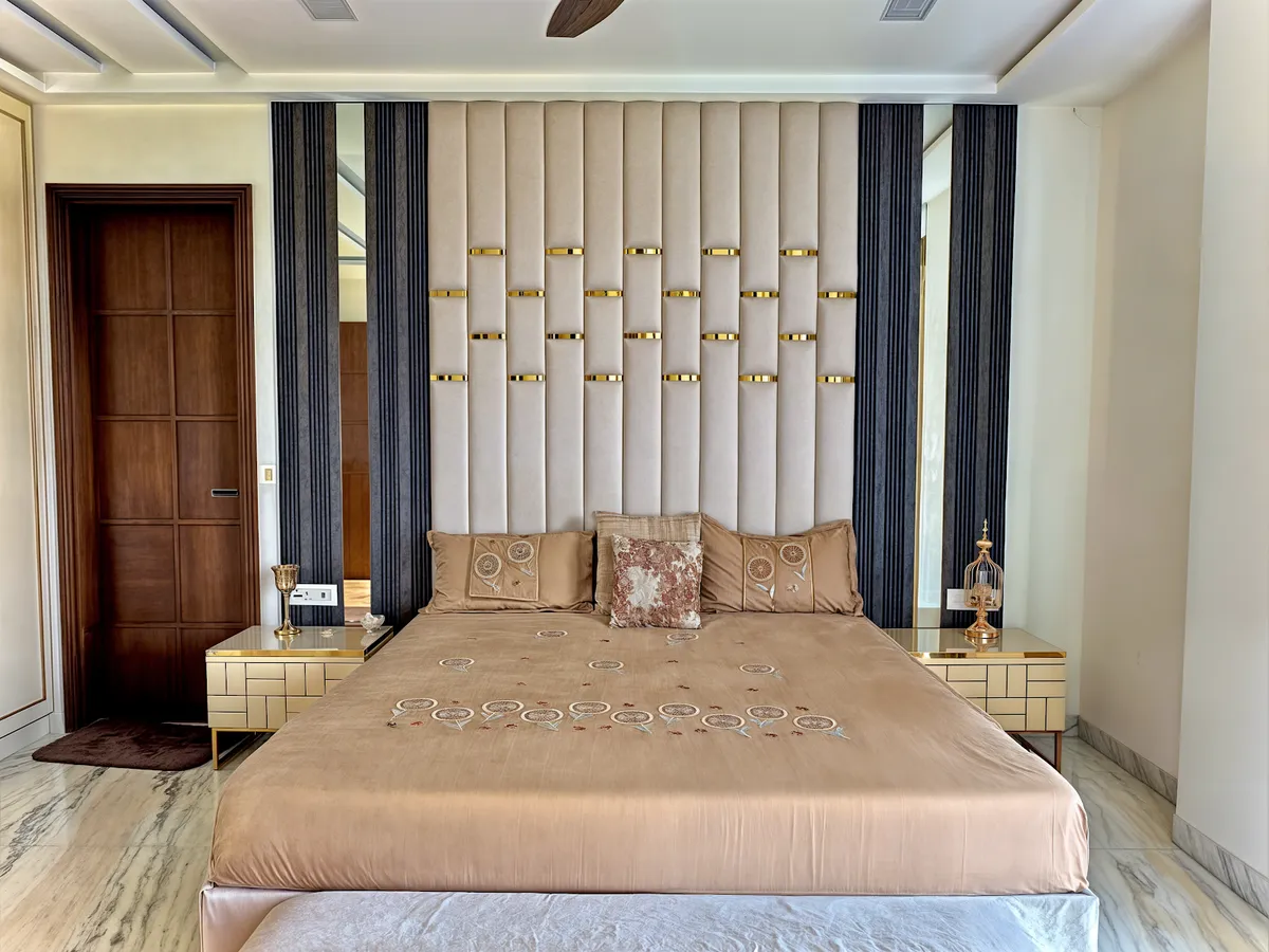 Symmetrical master bedroom with a full-height channel-tufted beige headboard wall studded with staggered brass straps