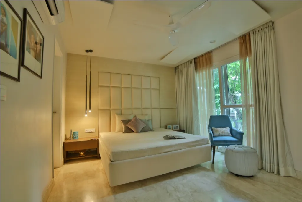 Cream bedroom with a grid-tufted upholstered headboard panel carried wall to wall, slim black cylinder pendants dropped over a floating bedside shelf