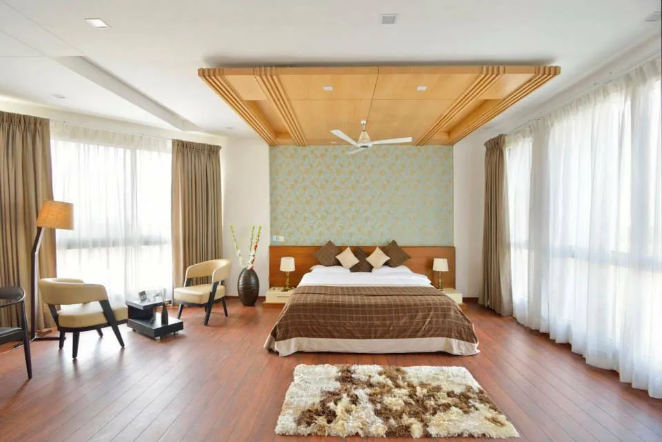 Spacious daylit master bedroom with a stepped maple-slat ceiling raft over the bed, a pale blue-and-gold damask wallpaper strip behind the wooden headboard