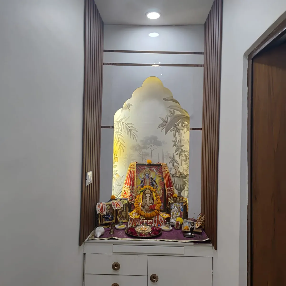 Built-in pooja niche framed by fluted wood columns with a backlit arched botanical glass panel, idols dressed in marigold garlands