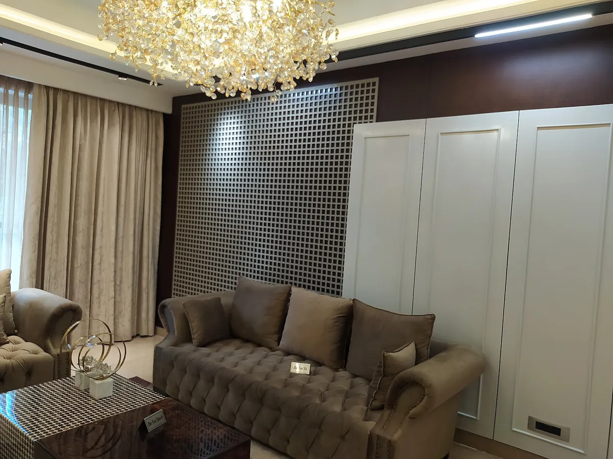 Show-flat living room: two deep-buttoned taupe velvet sofas carrying 'Do not sit' placards, facing a mirror-backed square lattice screen in a dark wood frame